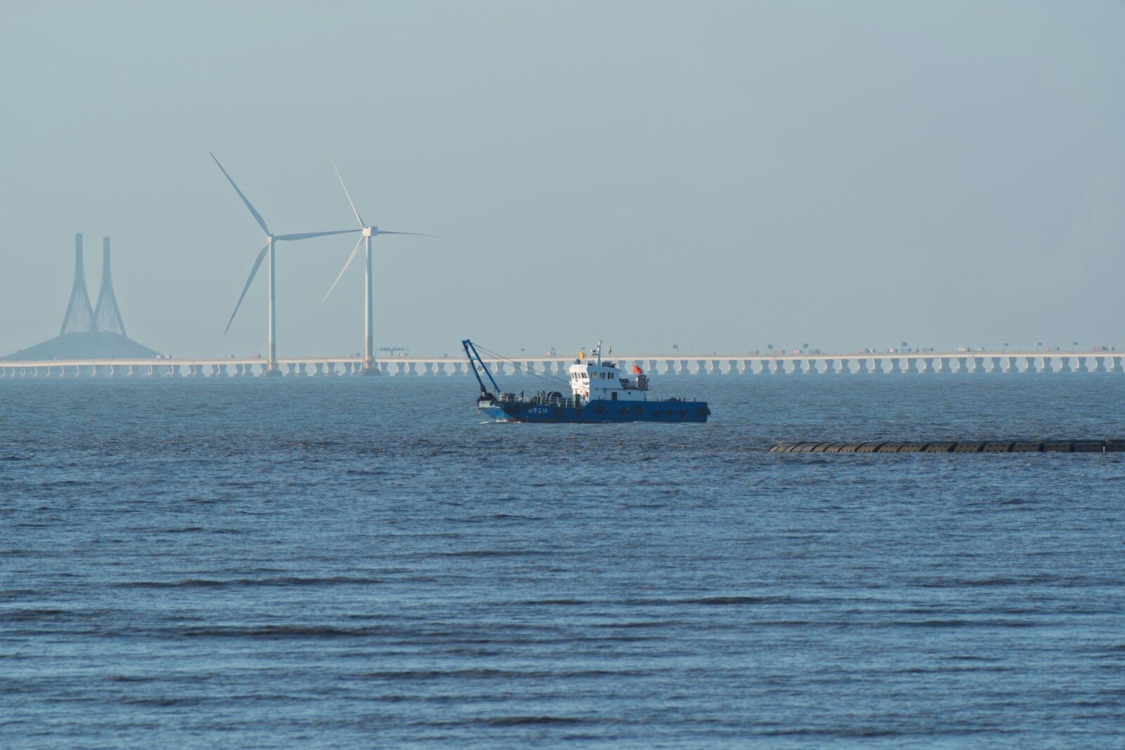A dredging boat sails past wind turbines and a bridge.